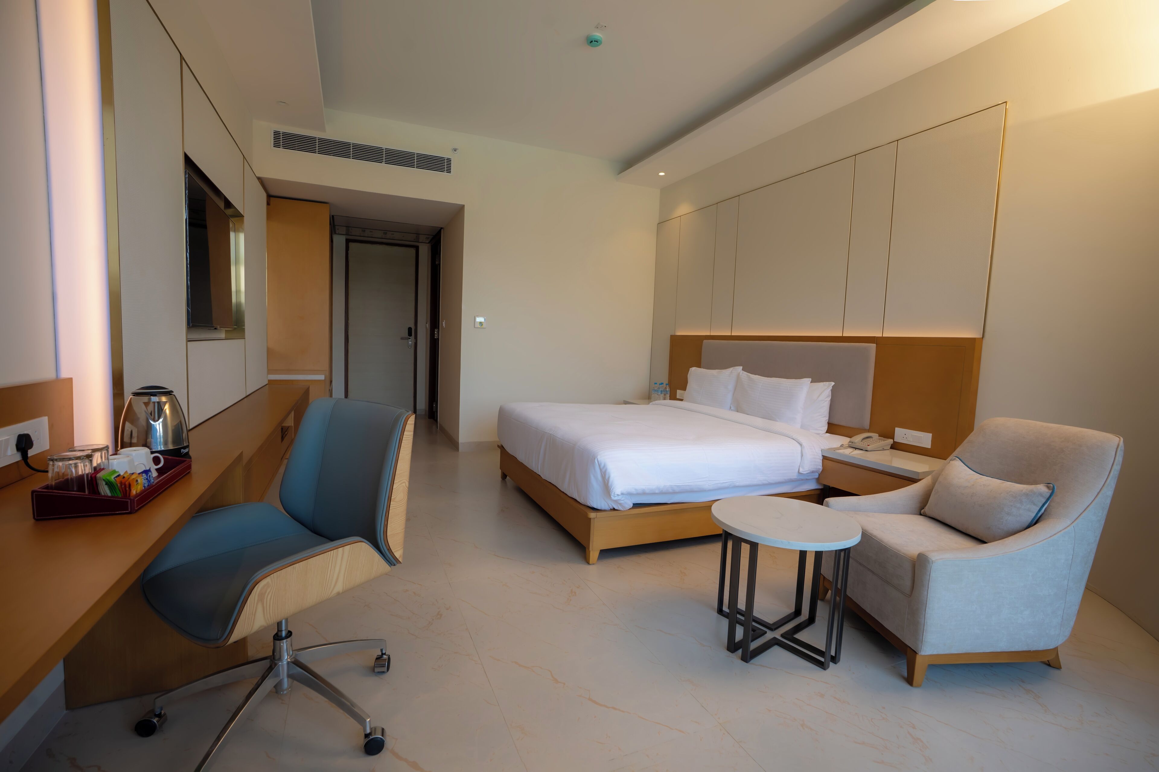 A modern hotel room with a bed, a desk with a coffee maker, a chair, and a small table.
