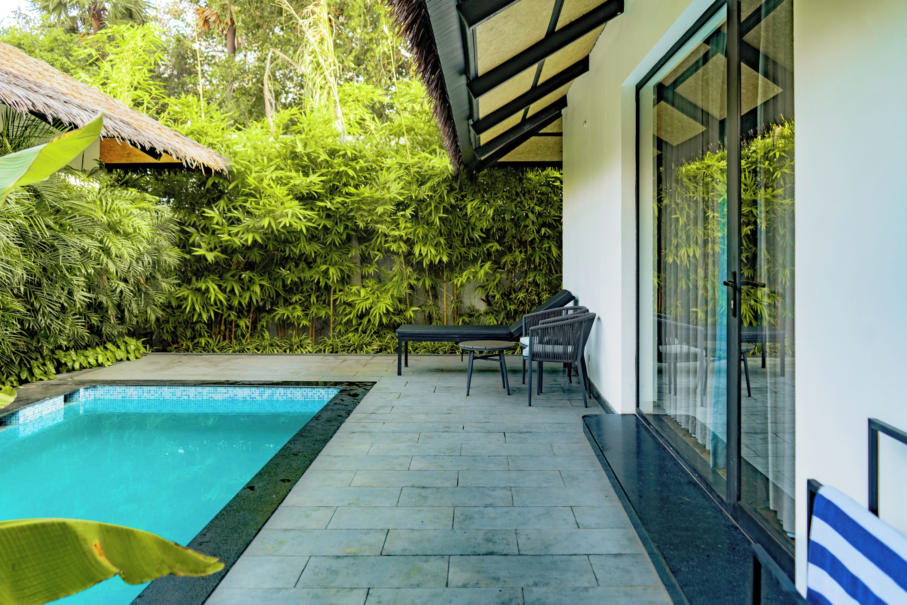 A swimming pool with a tiled deck, a covered seating area with chairs, and lush greenery surrounding the space.