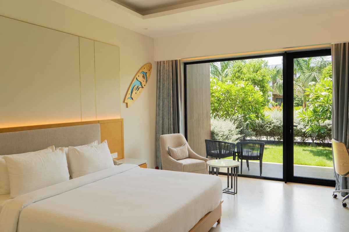 A hotel room with a large bed, a chair, a small table, and a view of greenery through the glass door.
