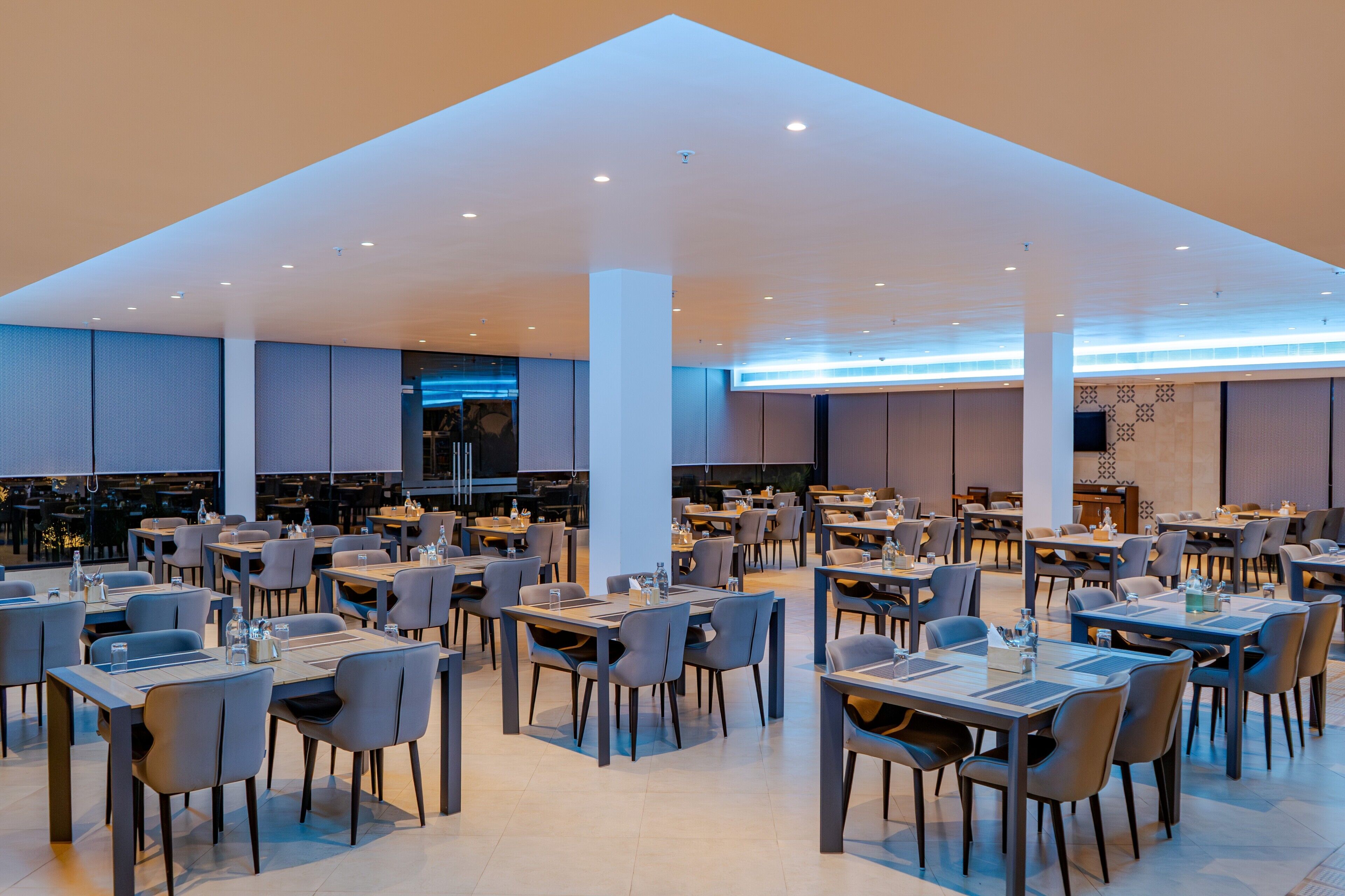 Un comedor amplio con varias mesas y sillas, un techo alto con iluminación empotrada y un diseño arquitectónico moderno.
