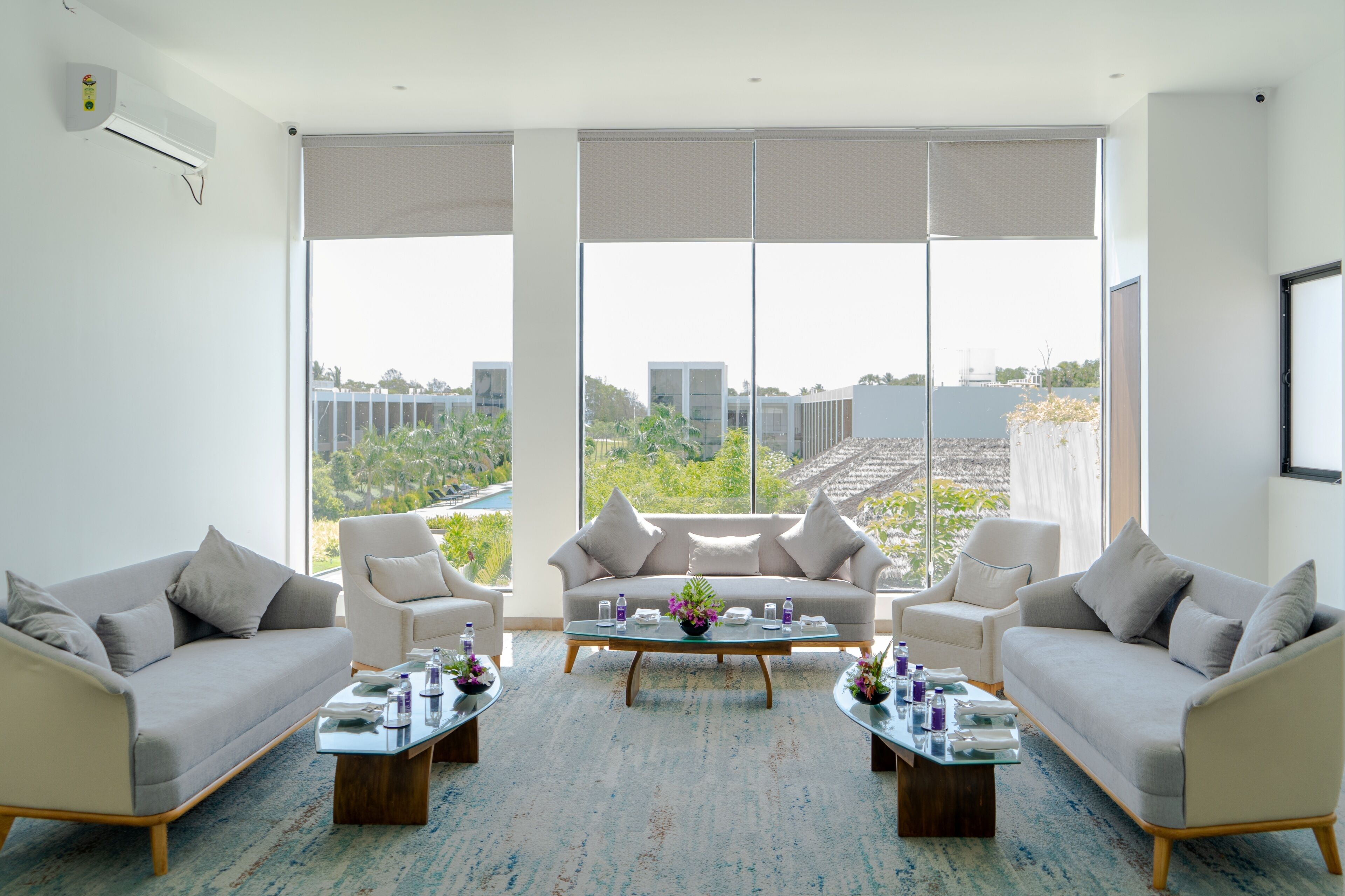 A modern living room with a sofa set, two coffee tables, and a large window with a view of greenery and buildings.