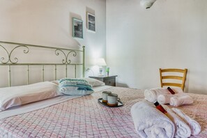 Uma cama bem arrumada com um cobre-leito estampado, uma mesinha de cabeceira com abajur e uma cadeira.