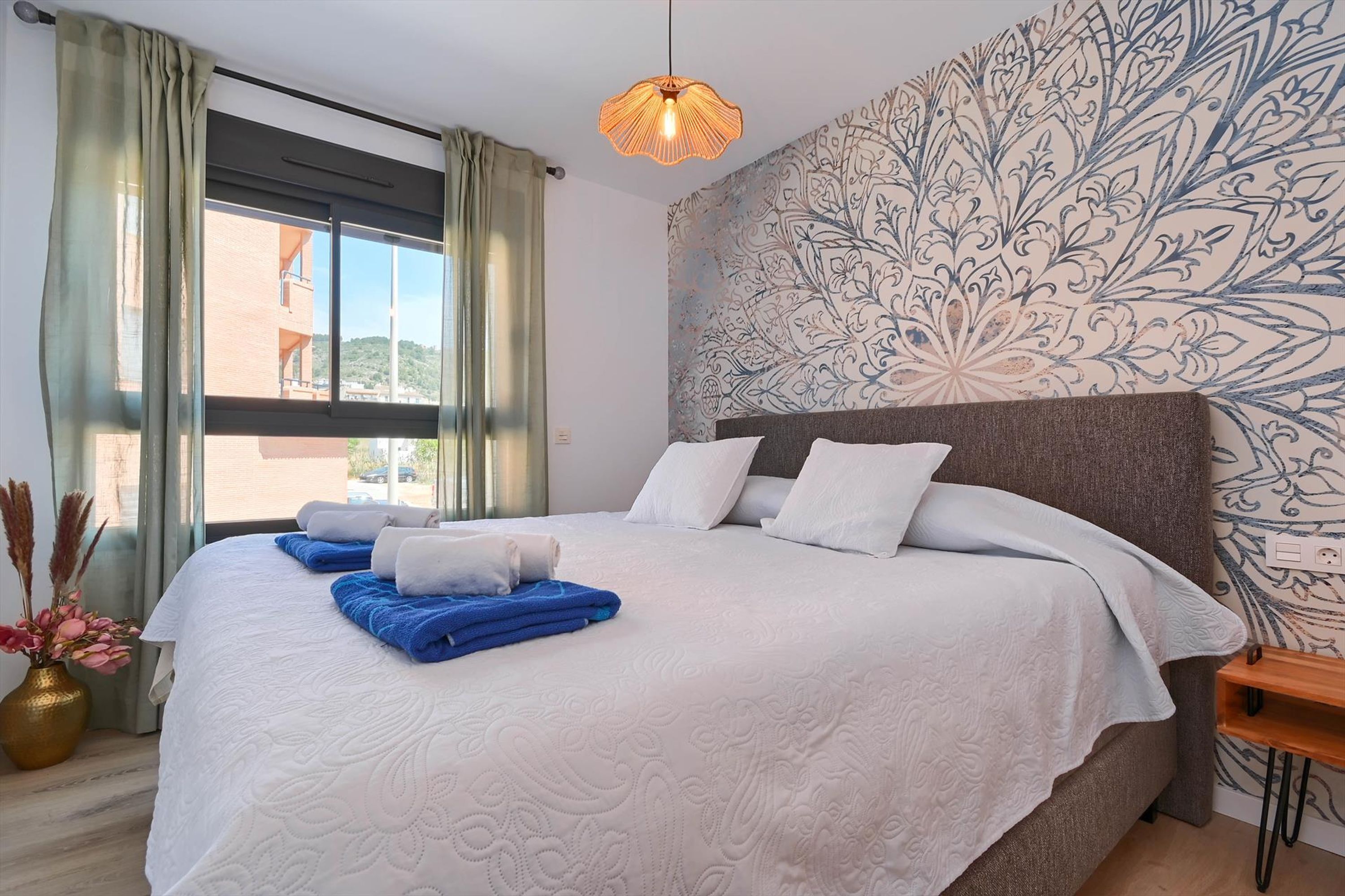 A neatly made bed with white and blue linens, a patterned wall, and a window with curtains.