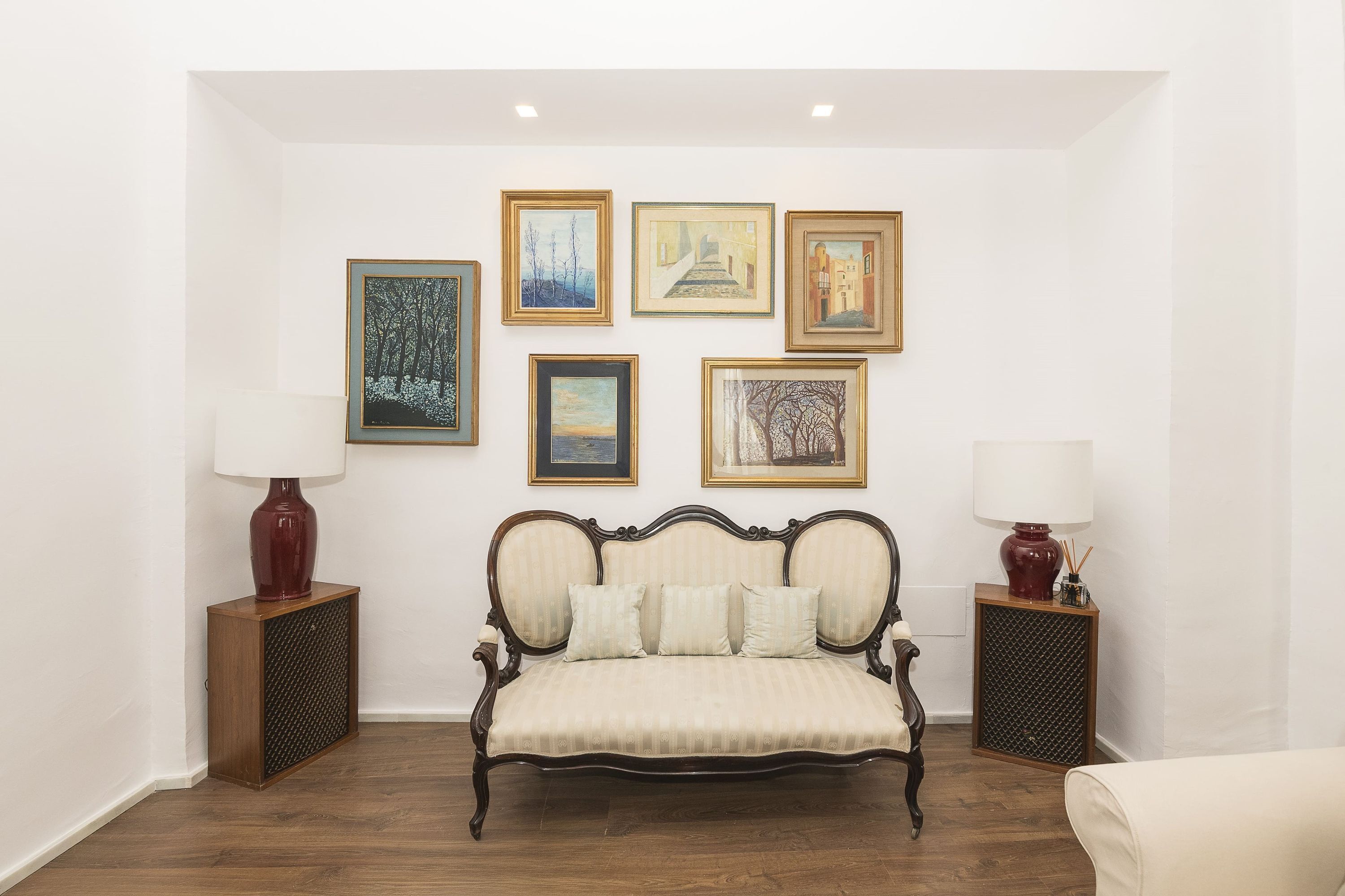 A room with a vintage-style sofa, two side tables with lamps, and six framed artworks on the wall.