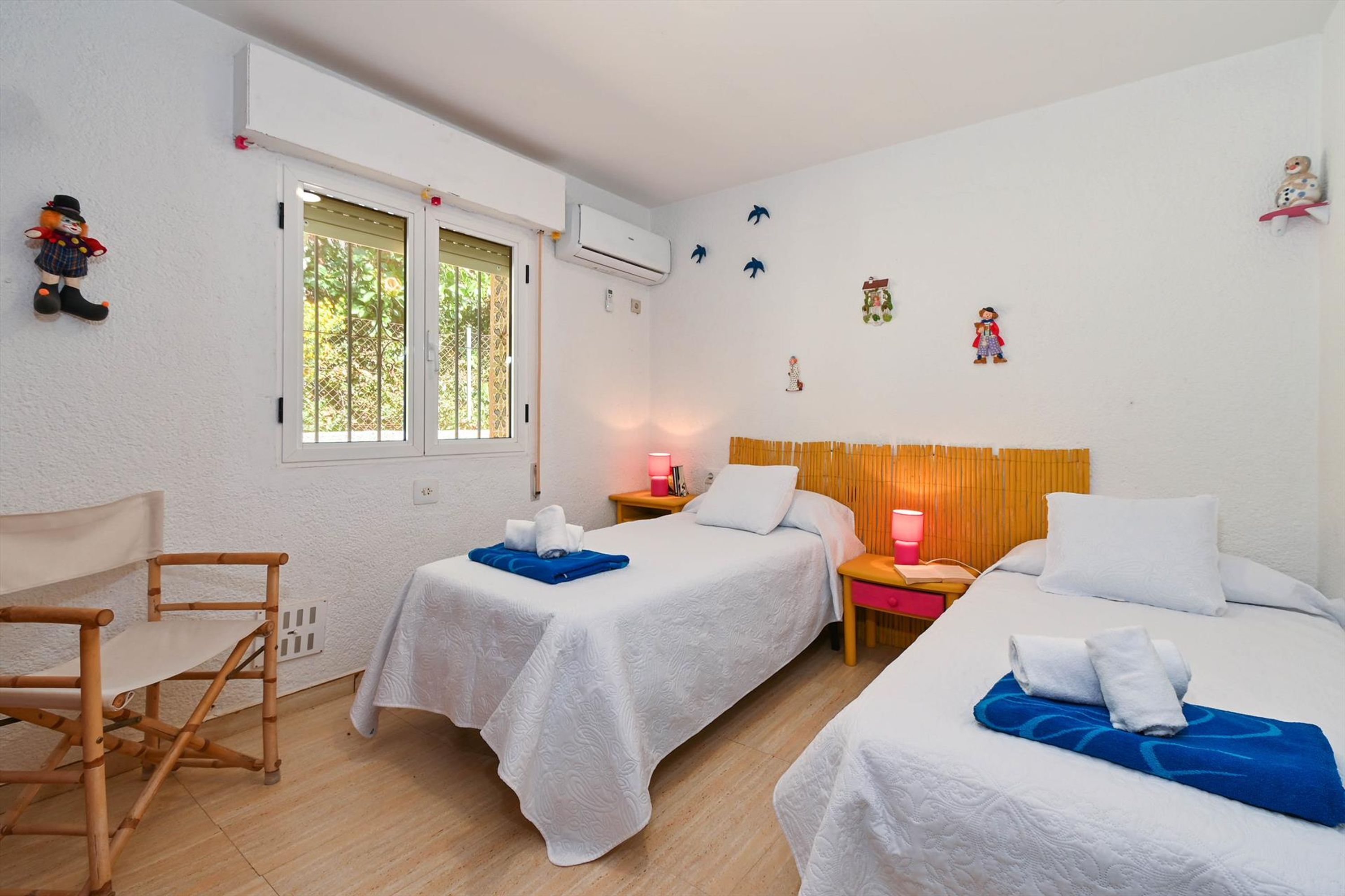 A room with two single beds, a small table, a chair, and a window with blinds.