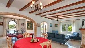 A spacious living area with a red-covered dining table, wooden chairs, a blue sofa, and a chandelier.