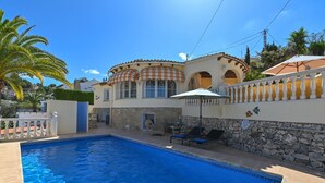 Μια πισίνα με ξαπλώστρες και ομπρέλες, ένας πέτρινος τοίχος και ένα κτίριο με τοξωτά παράθυρα και κεραμοσκεπή.