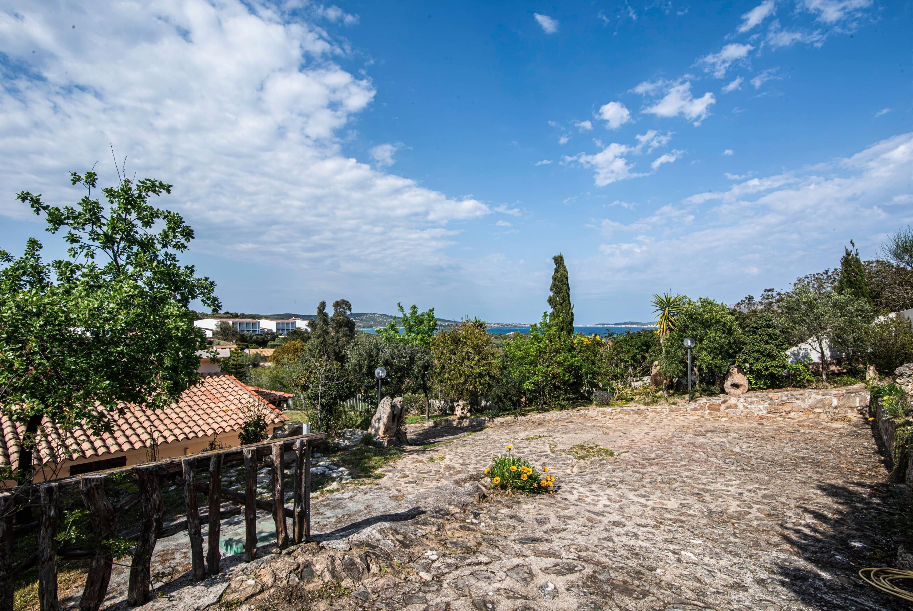 Une cour pavée de pierres, avec un bâtiment surmonté d’un toit de tuiles, entouré d’arbres et sous un ciel dégagé.