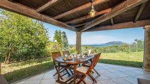 An outdoor patio with a wooden table and chairs, a hanging light fixture, and a view of a mountainous landscape. - Gli Allori in Golfo Aranci (Golfo Aranci)