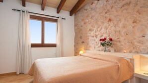 Un dormitorio con pared de piedra, una cama con cubrecama color beige, una ventana con cortinas y una mesita de noche con lámpara y flores.
