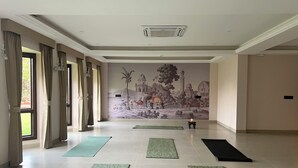 A spacious room with large windows, a mural depicting a historical scene, and several yoga mats on the floor. - Kyra Wellness Resort (Hyderabad)