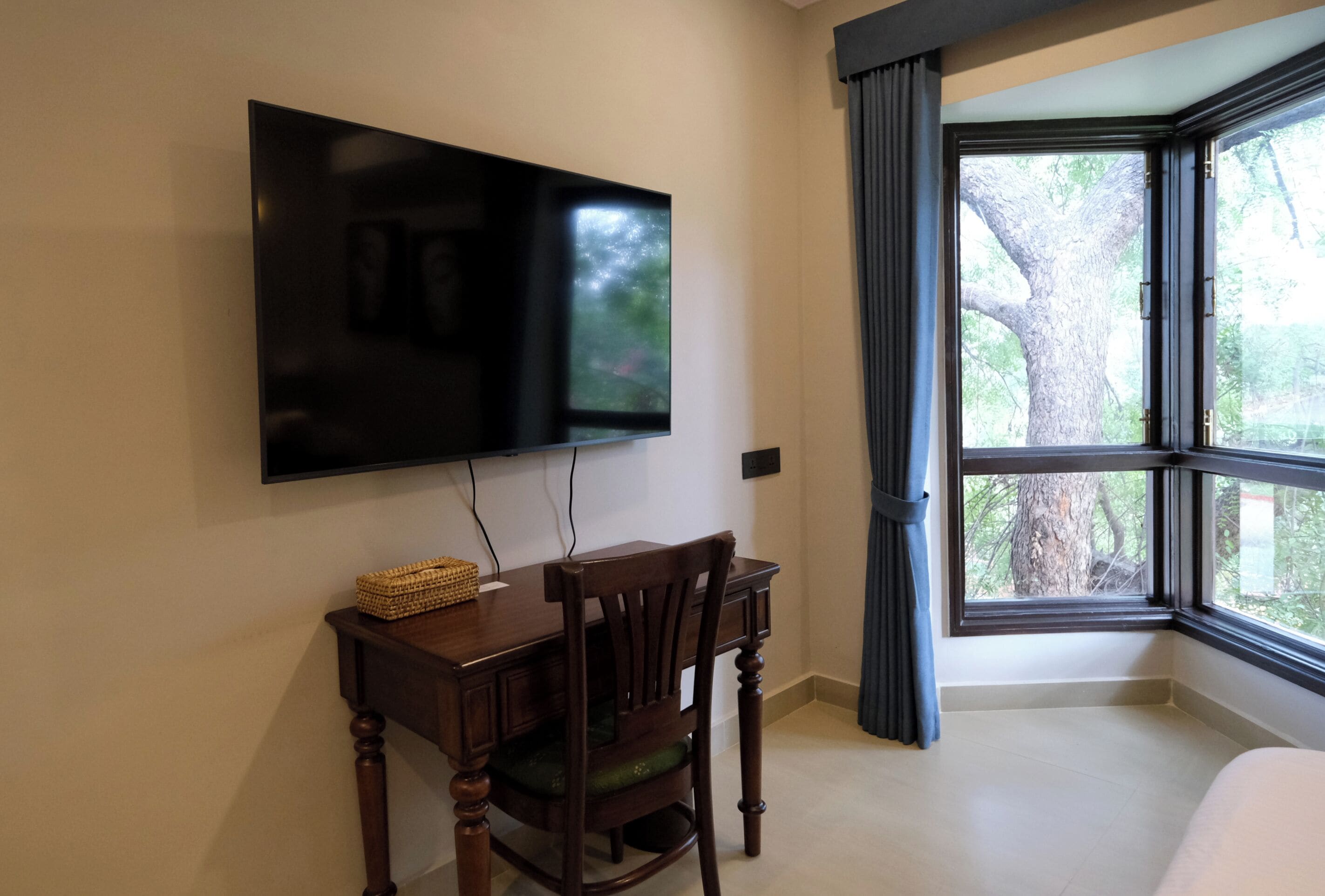 A room with a flat-screen TV mounted on the wall, a wooden desk with a chair, and a large window with curtains.