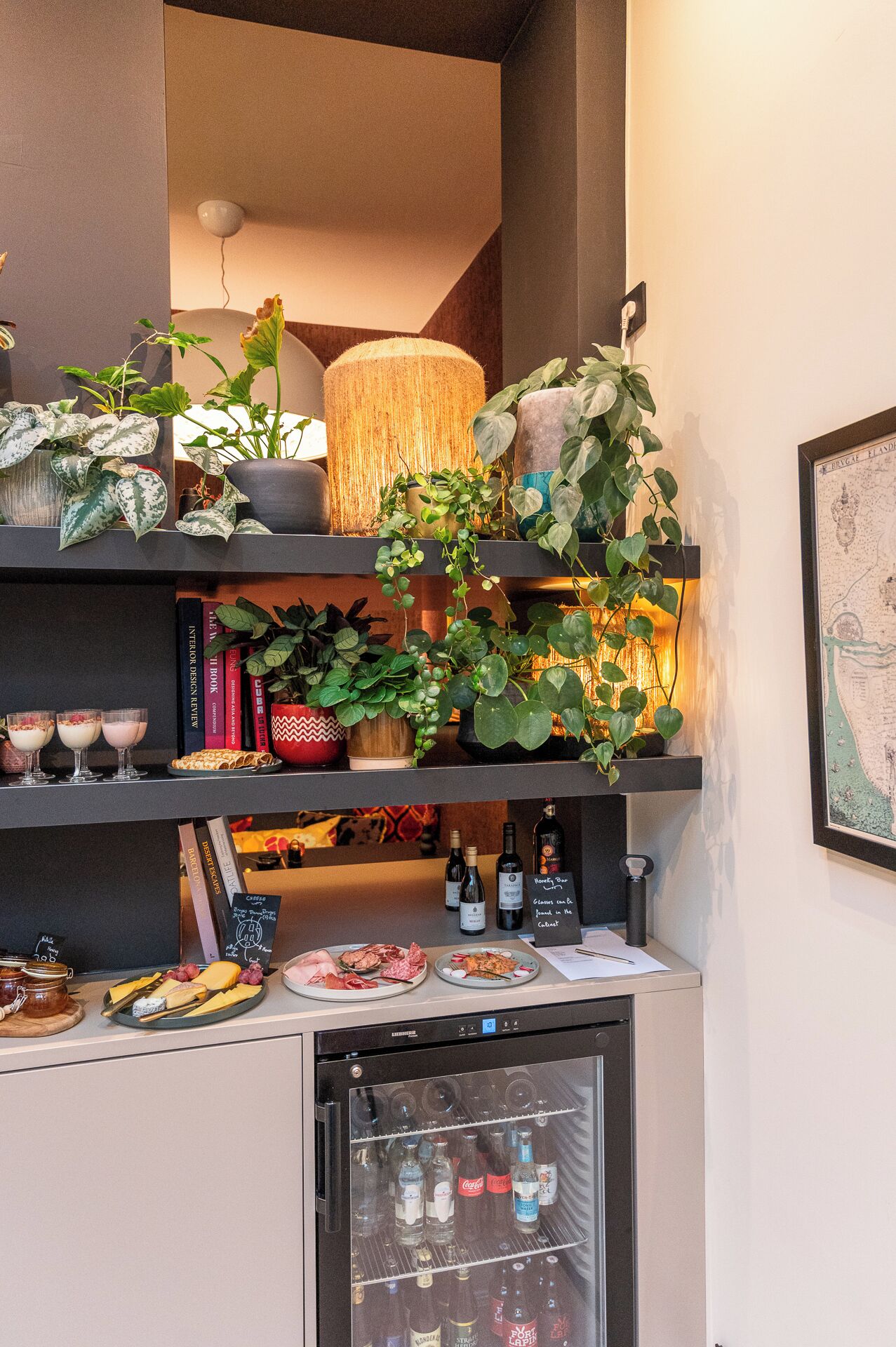 Una zona de cocina con estantes que exhiben plantas, libros y alimentos. Se observa un refrigerador con botellas y una pequeña cafetera.