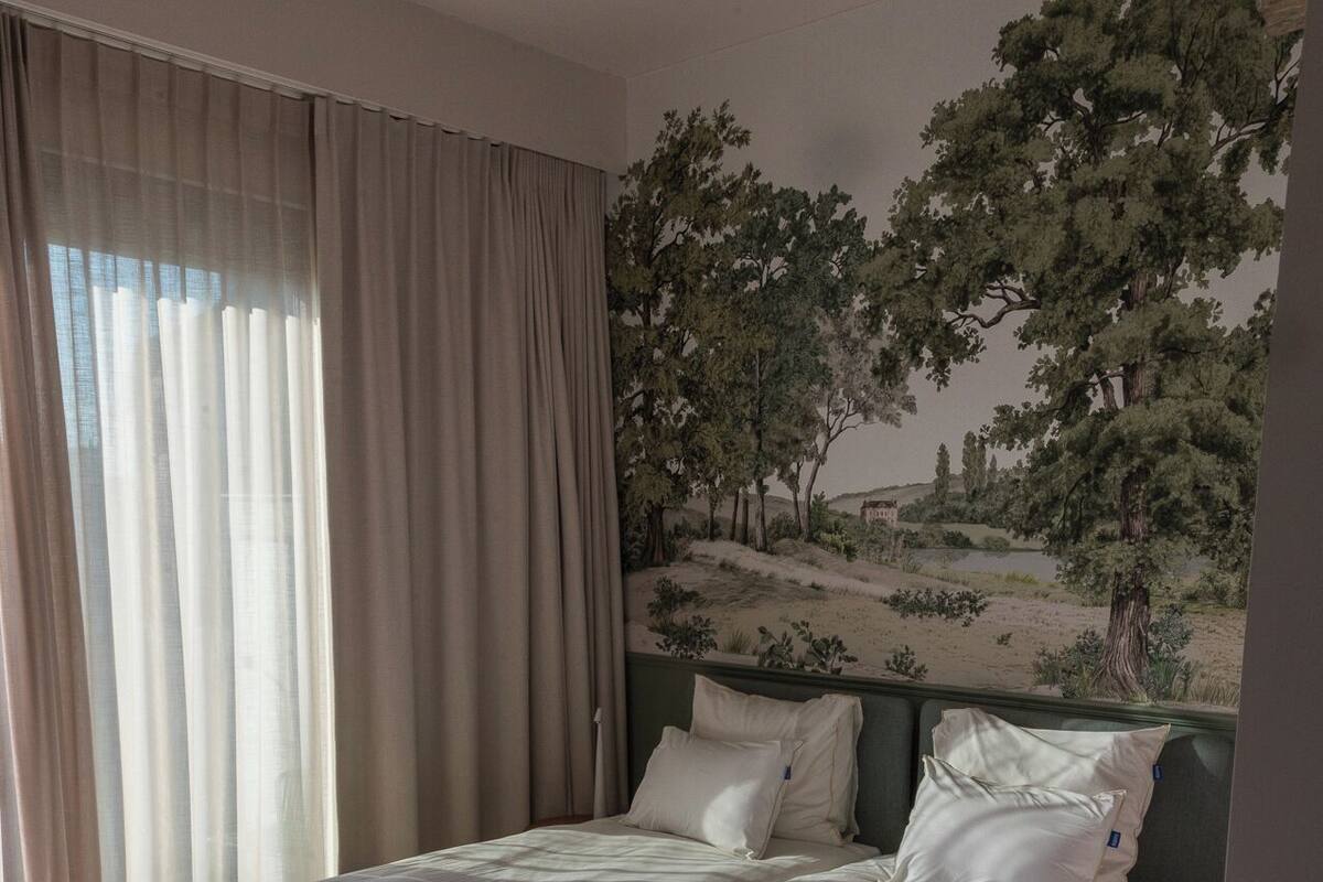 A bedroom with a bed, curtains, and a mural of a landscape.