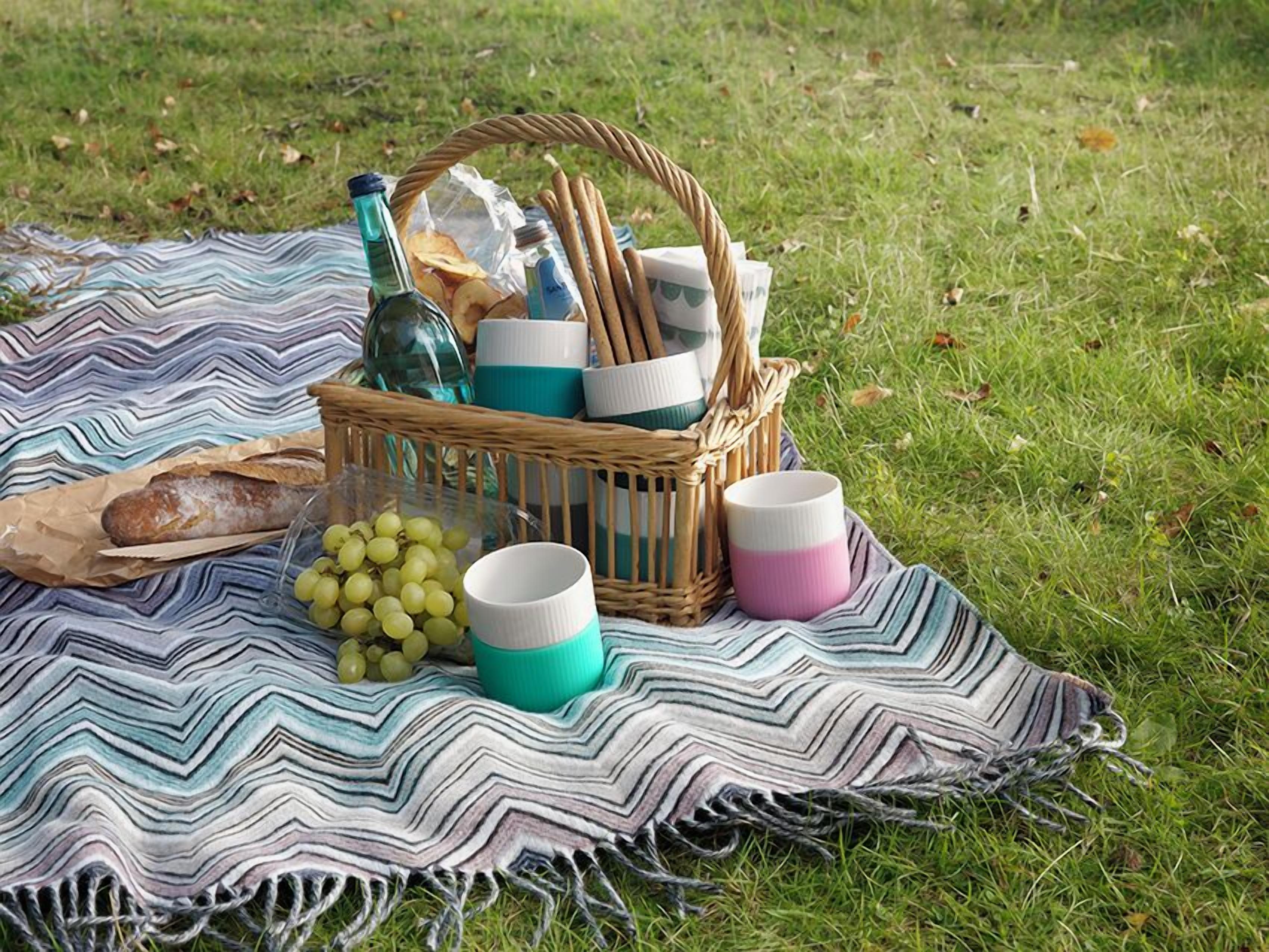 Un montaje para picnic en un área verde con una manta a rayas, una cesta de mimbre que contiene pan, una botella y vasos.