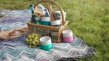 Un montaje para picnic en un área verde con una manta a rayas, una cesta de mimbre que contiene pan, una botella y vasos.