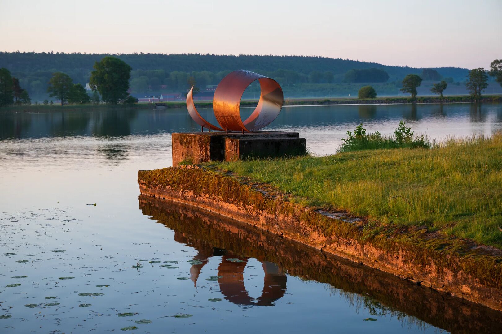 Una escultura metálica frente a un lago tranquilo con una orilla cubierta de hierba y árboles al fondo.