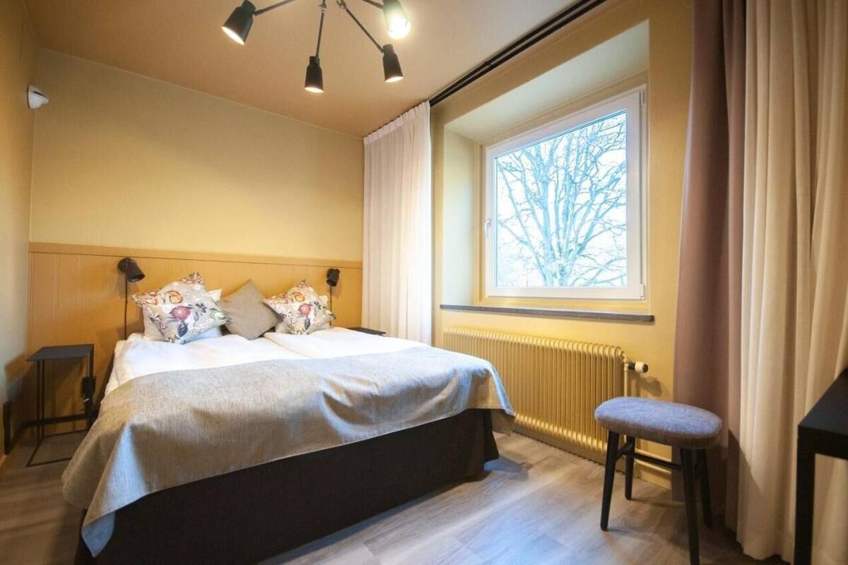 Une chambre à coucher avec un lit, une fenêtre avec des rideaux, un radiateur et un petit tabouret.