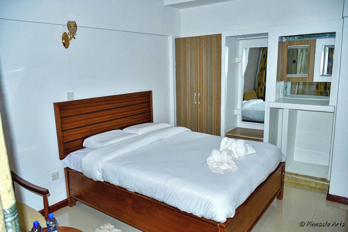 A neatly made bed with white linens, a wooden headboard, and a wooden wardrobe with a mirror above it.