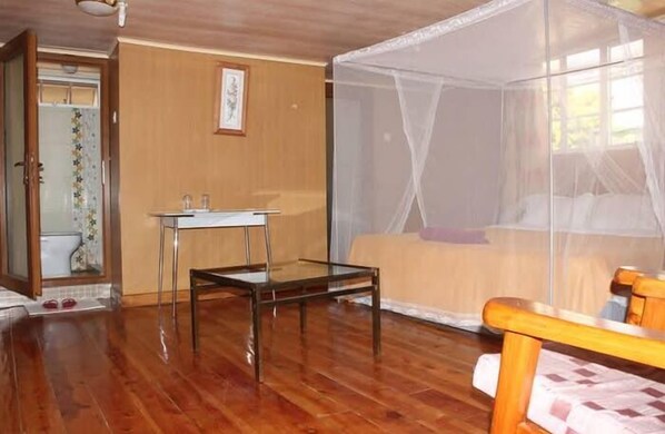 A room with a wooden floor, a bed with a mosquito net, a small table, and a chair. - Classic Guest Houss (Nairobi)