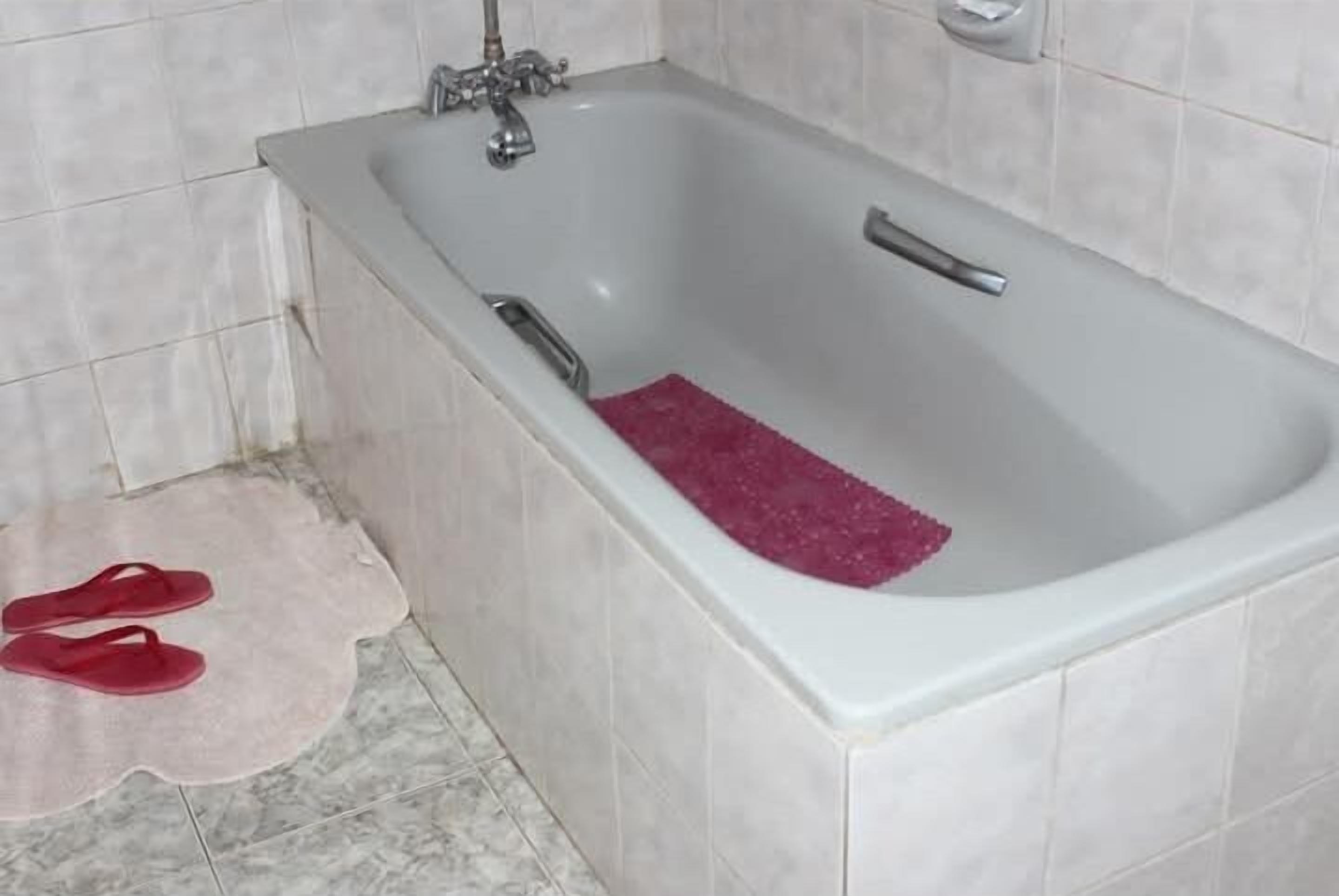 A bathroom with a bathtub, a red bath mat, and a pair of red slippers on the floor.