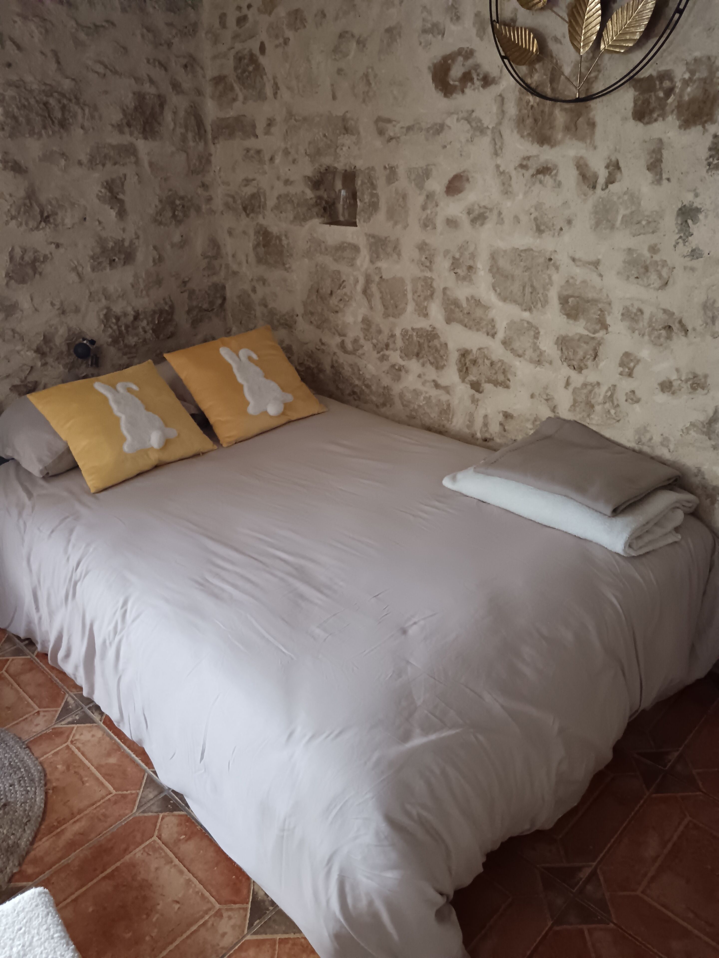Cama com roupa de cama branca, dois travesseiros amarelos com desenhos de coelhos brancos, uma luminária pequena na parede e uma parede de pedra.