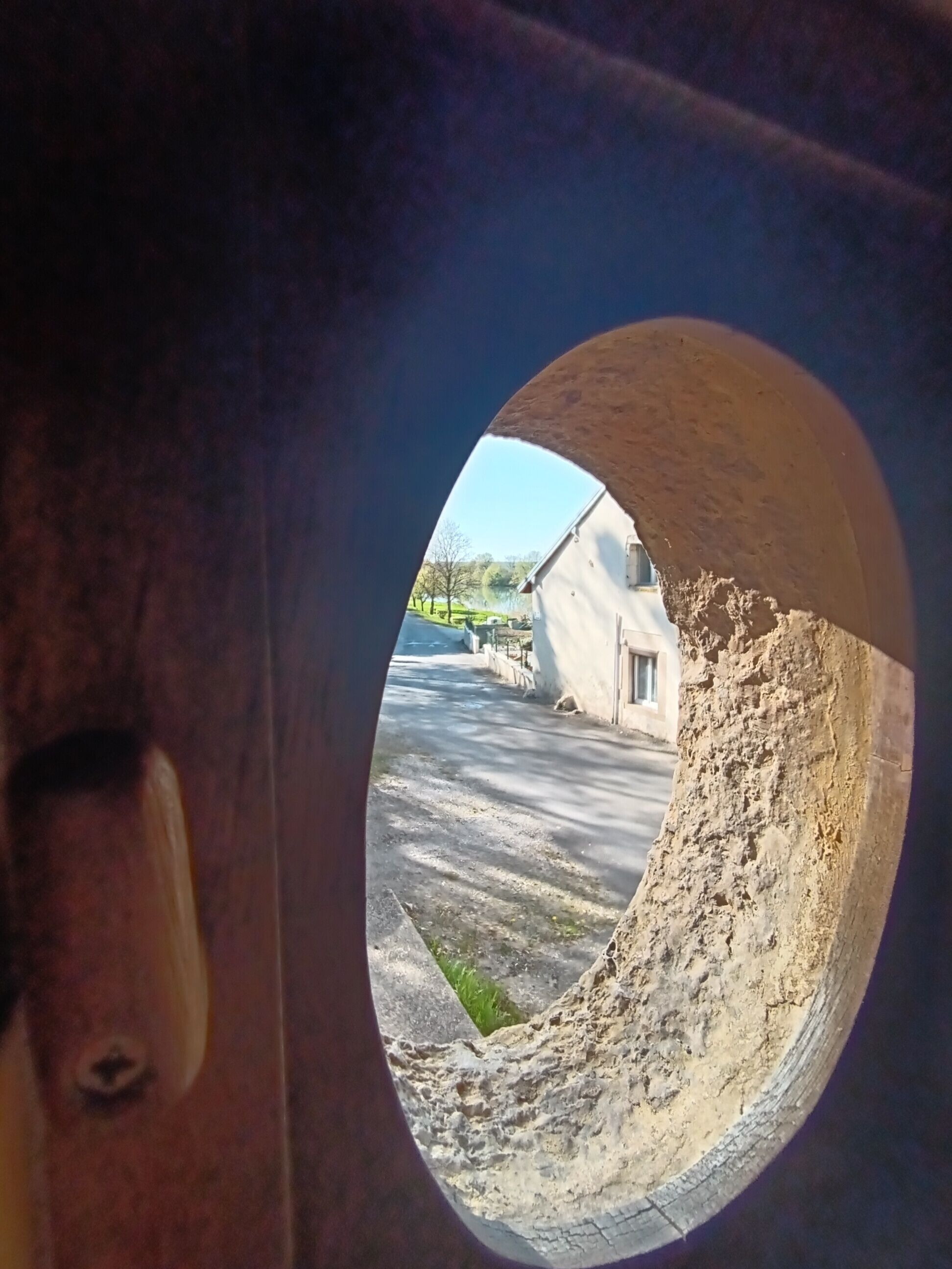 A view through a circular window of a building with a white exterior, a paved area in front, and a clear sky.