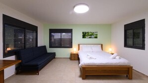 A bedroom with a bed, a sofa, two nightstands with lamps, and a window with blinds. - Adi Hacarmel (Daliyat al-Karmel)