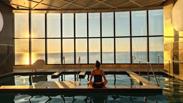 A person meditating by a large indoor pool with a view of the sunset.