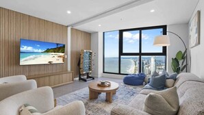 A modern living room with a large TV, comfortable seating, a coffee table, and a view of the city through large windows.