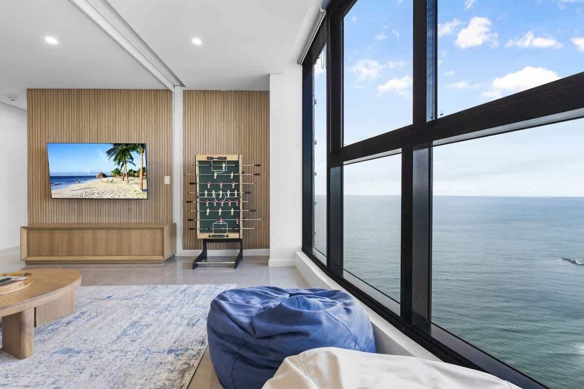 A room with a large window overlooking the sea, a foosball table, and a television displaying a beach scene.