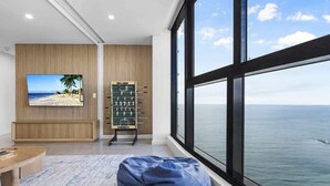 A room with a large window overlooking the sea, a foosball table, and a television displaying a beach scene.