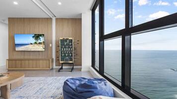 A room with a large window overlooking the sea, a foosball table, and a television displaying a beach scene.