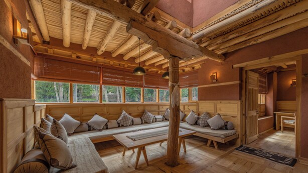 A cozy log cabin interior with a long bench seating area, wooden beams, and large windows. - Chulli Bagh Villa  (Leh)