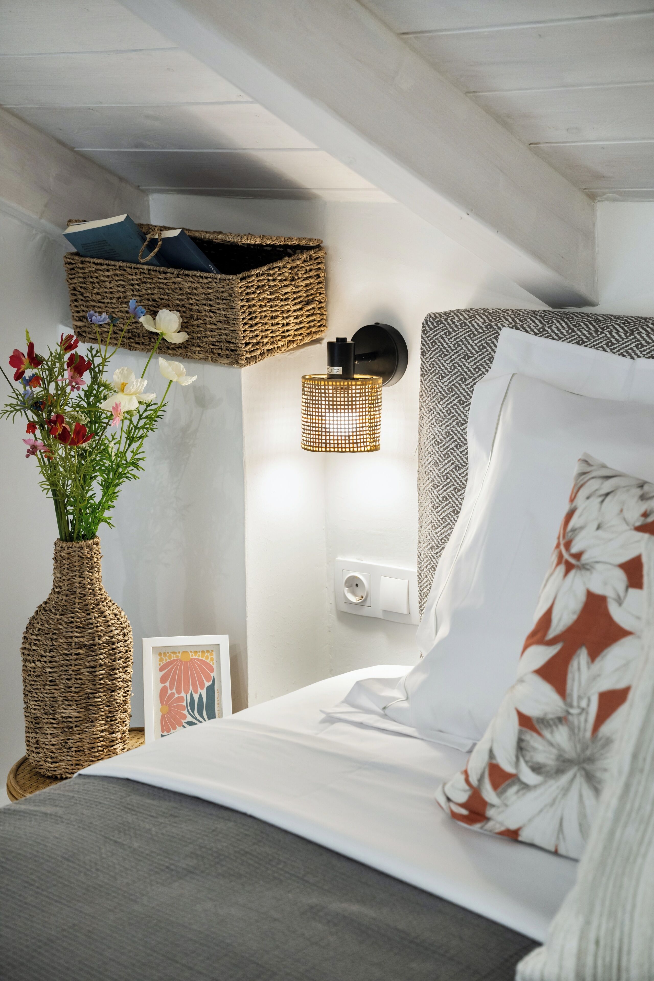 A bedroom with a bed, a wicker basket, a wall-mounted lamp, and a vase with flowers.