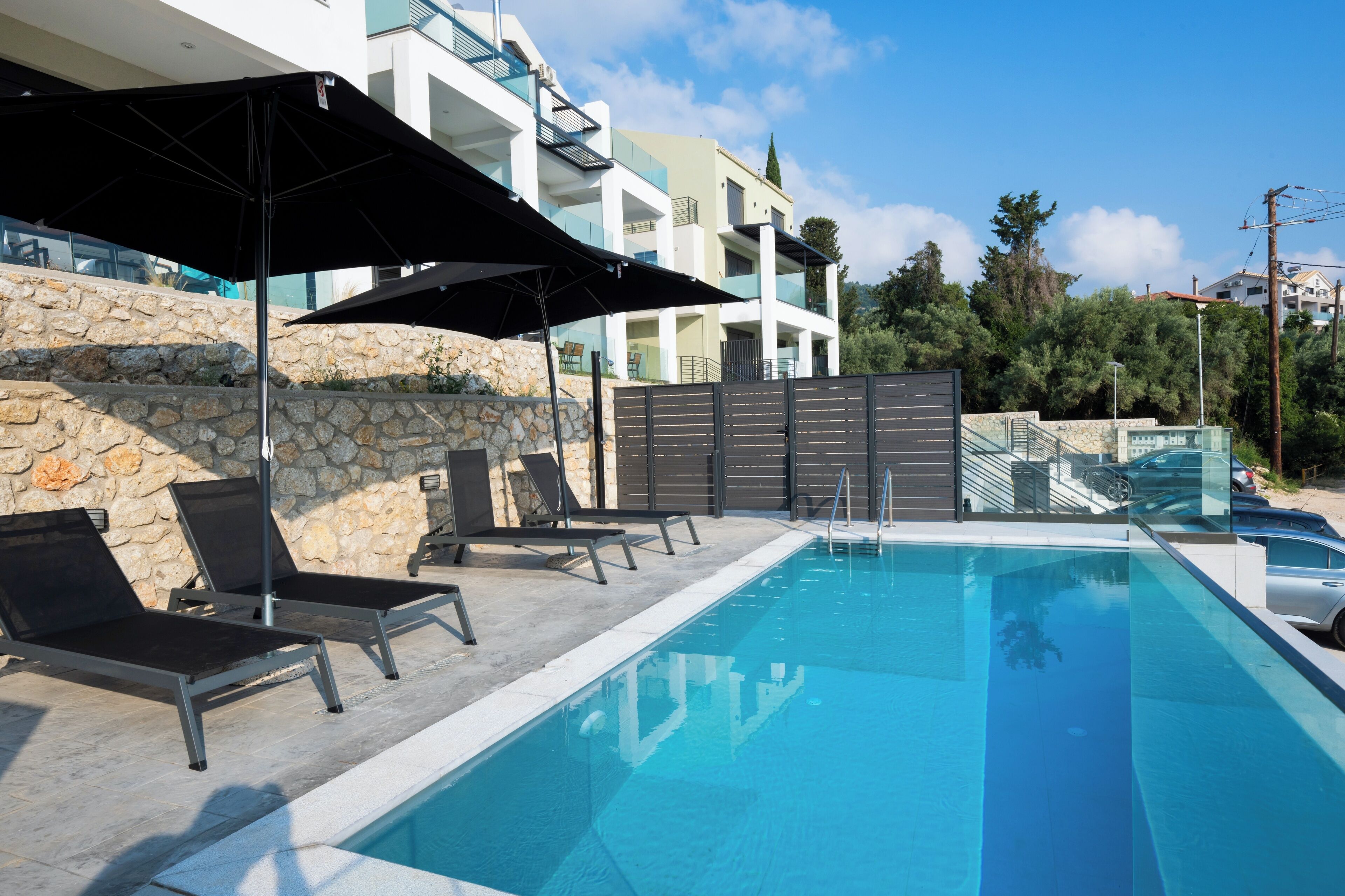 A swimming pool with lounge chairs and umbrellas, surrounded by stone walls and modern buildings.