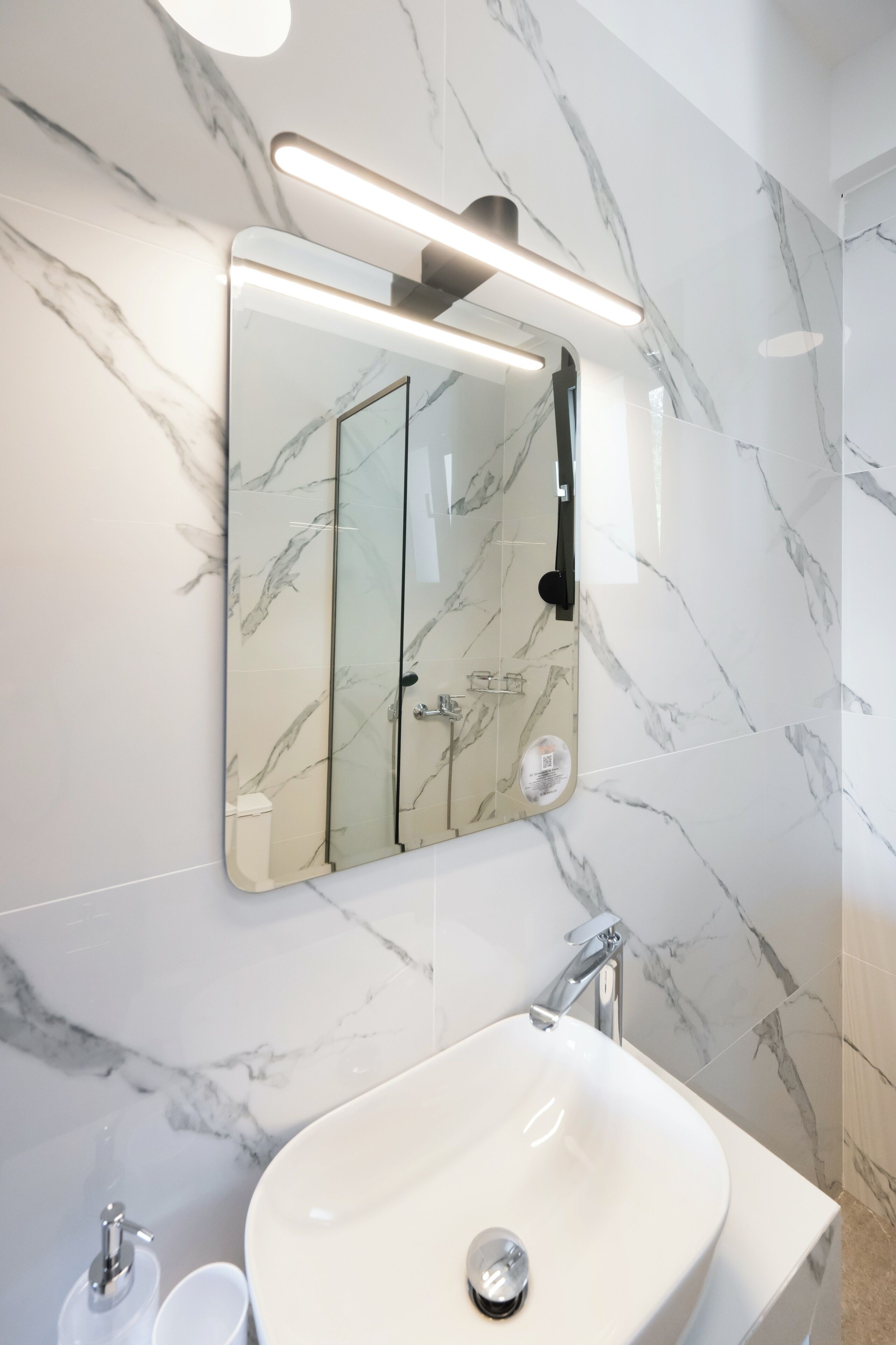 Un baño con pared de mármol, un lavabo blanco y un espejo con luz superior.