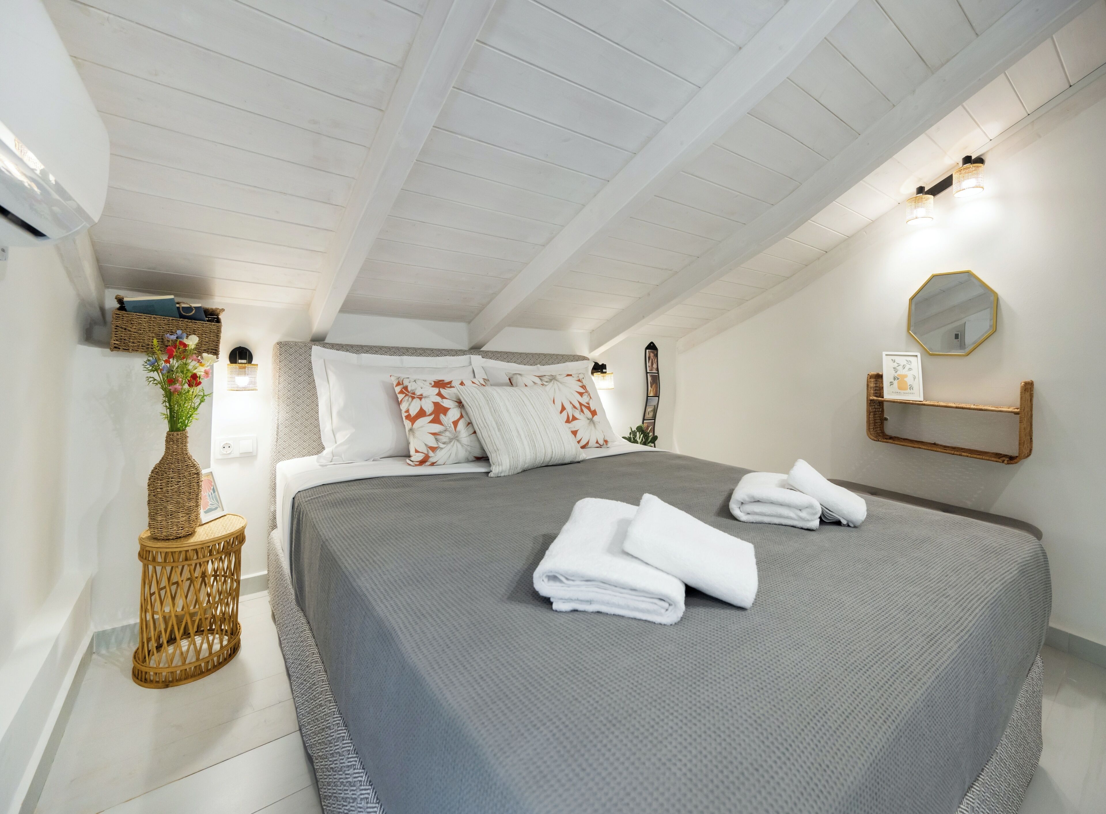 Une chambre avec un grand lit, des murs blancs, un plafond en bois et une petite étagère sur laquelle se trouvent un miroir et un cadre photo.