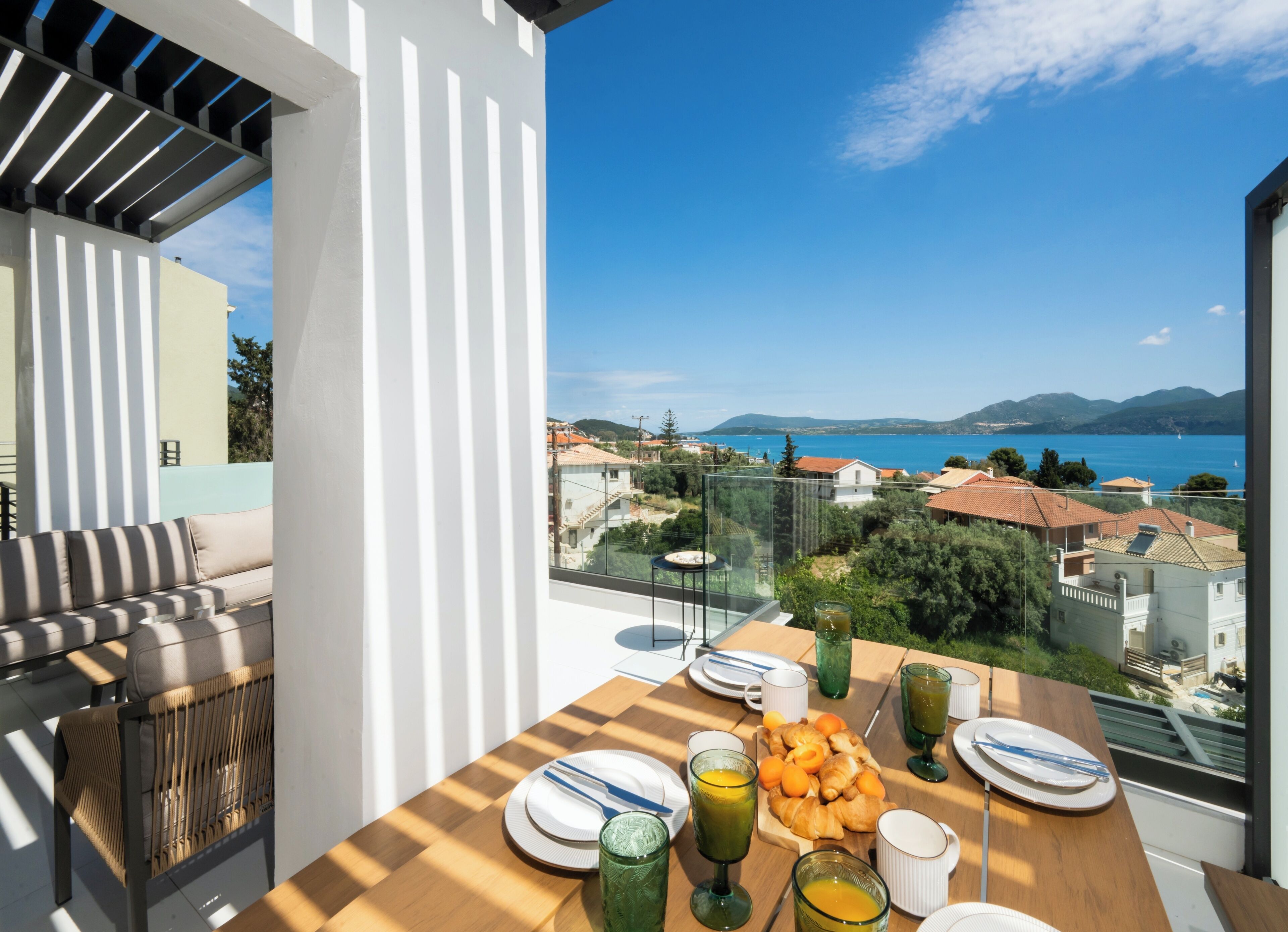 Un balcone con un tavolo di legno apparecchiato per la colazione, con vista panoramica su una cittadina costiera e sul mare.