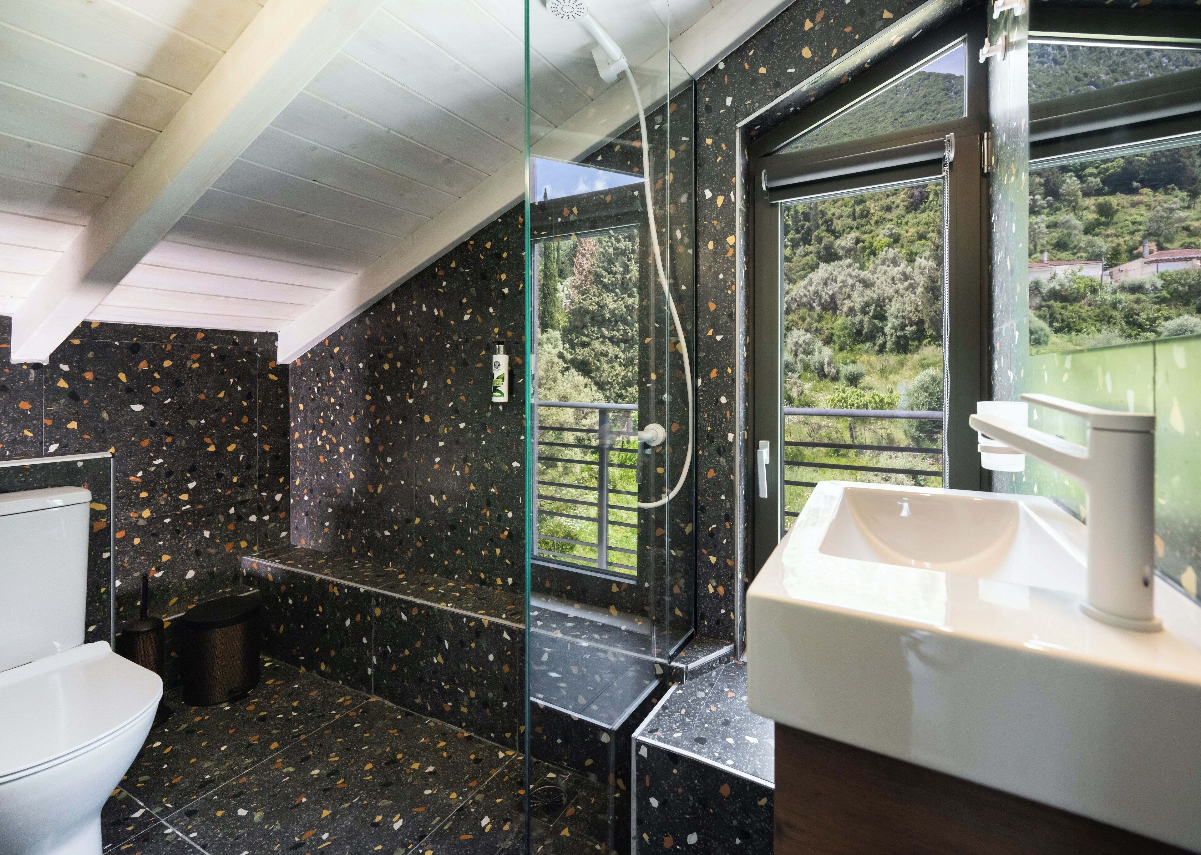 Un baño con pared de baldosas negras con motas, un lavamanos blanco y una ventana que da a zonas verdes.
