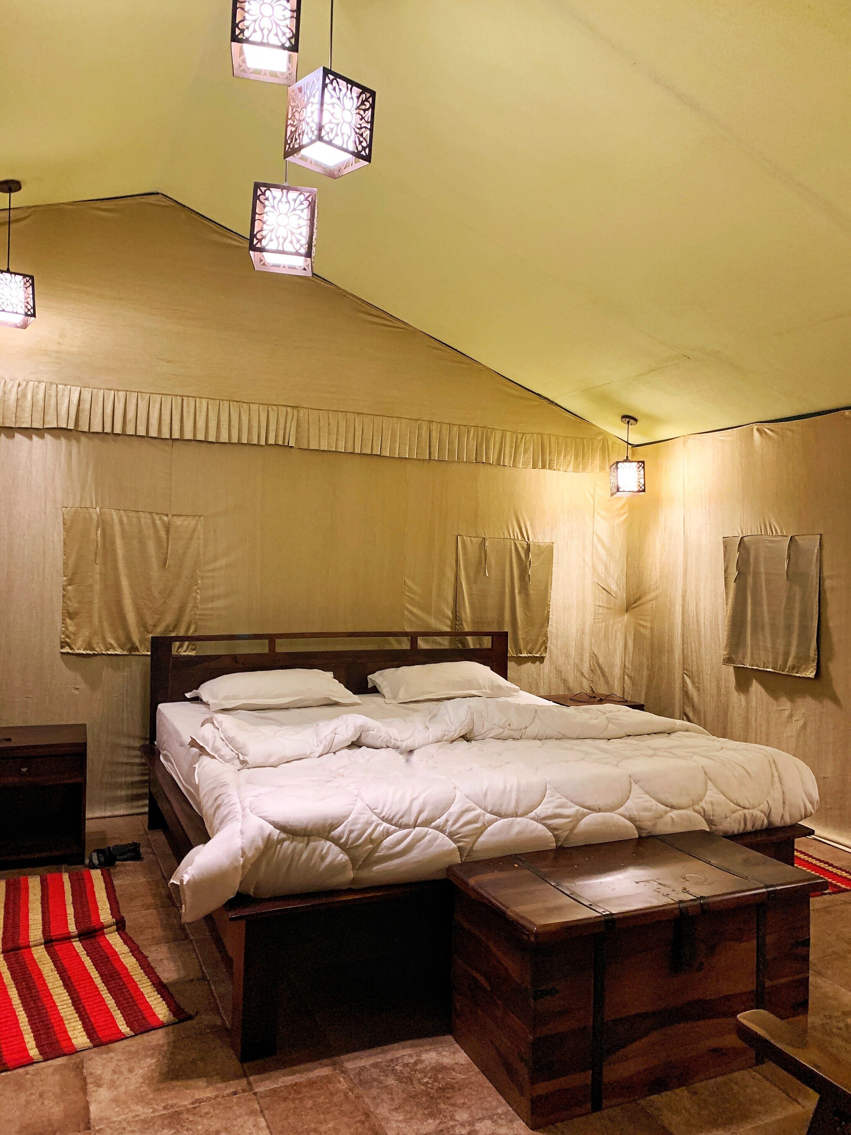 A bedroom with a large bed, wooden headboard, and a nightstand. There are decorative hanging lights and a red and yellow striped rug on the floor.