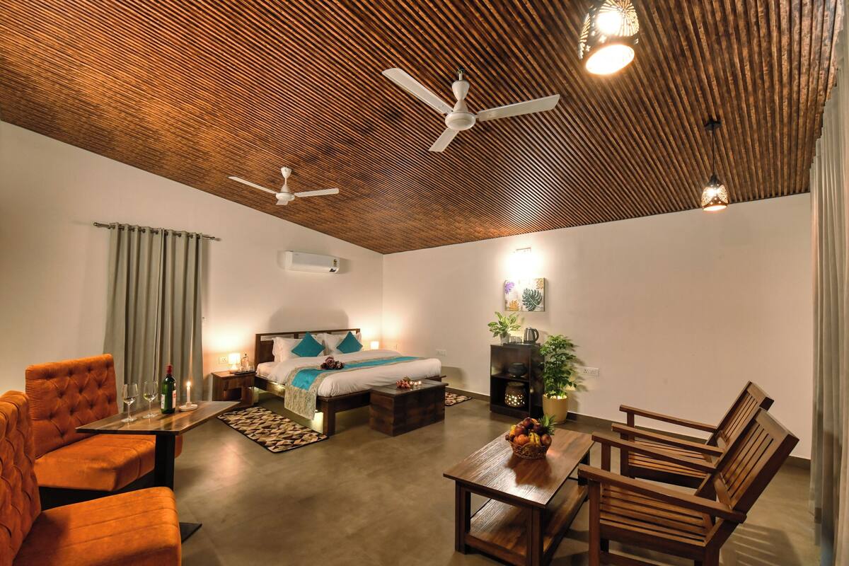 A modern room with a wooden ceiling, a bed with a white headboard, a seating area with orange chairs, a wooden table with fruit, and a ceiling fan.