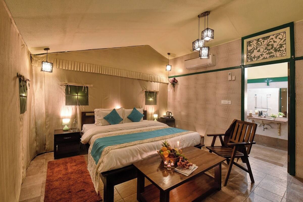 A hotel room with a bed, bedside tables, a desk with a chair, a small table with fruit and candles, and a bathroom visible through a glass door.