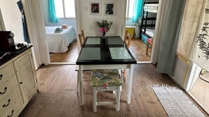 Une pièce comprenant une table à manger, des chaises, un téléviseur fixé au mur, un lit avec une tête de lit et une fenêtre avec des rideaux.