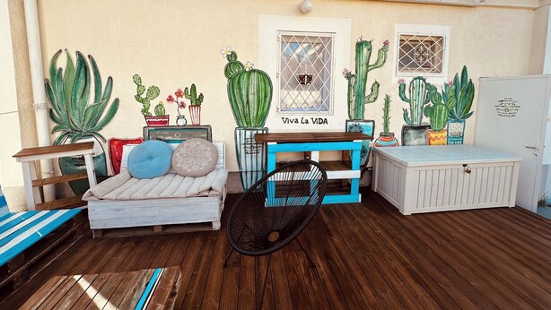 Un espace patio doté d’une terrasse en bois, d’un canapé blanc avec des coussins, d’une chaise bleue et de différents cactus en pot.