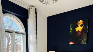 Een slaapkamer met een bed, nachtkastje, lamp, schilderij en uitzicht door een raam.