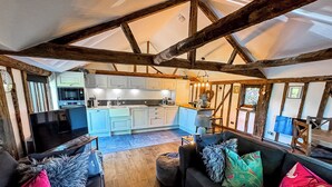 A spacious living area with a dark sofa set, a kitchen in the background, and exposed wooden beams. - Historic Luxury 18th C Listed Barn Apartment (Maresfield)