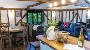 A rustic interior with exposed wooden beams, a wooden dining table with chairs, a sofa with cushions, a coffee table with a pitcher and basket, and a bottle of wine on the counter. - Historic Luxury 18th C Listed Barn Apartment (Maresfield)