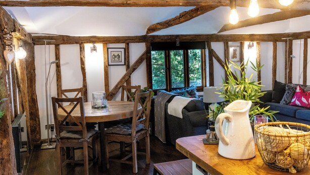 A rustic interior with exposed wooden beams, a wooden dining table with chairs, a sofa, and a coffee table with a pitcher and basket. - Historic Luxury 18th C Listed Barn Apartment (Maresfield)