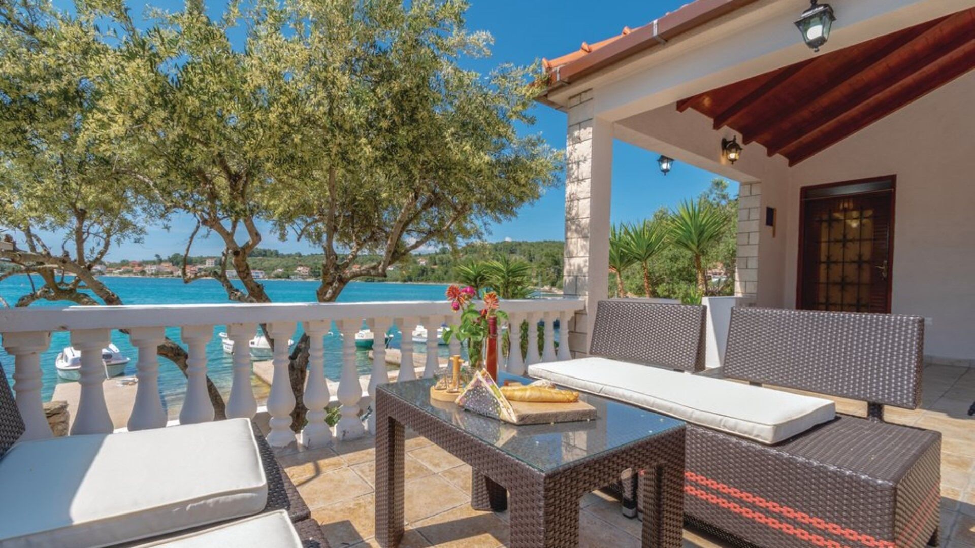 A terrace with outdoor seating, a view of the sea, and a building with a tiled roof.
